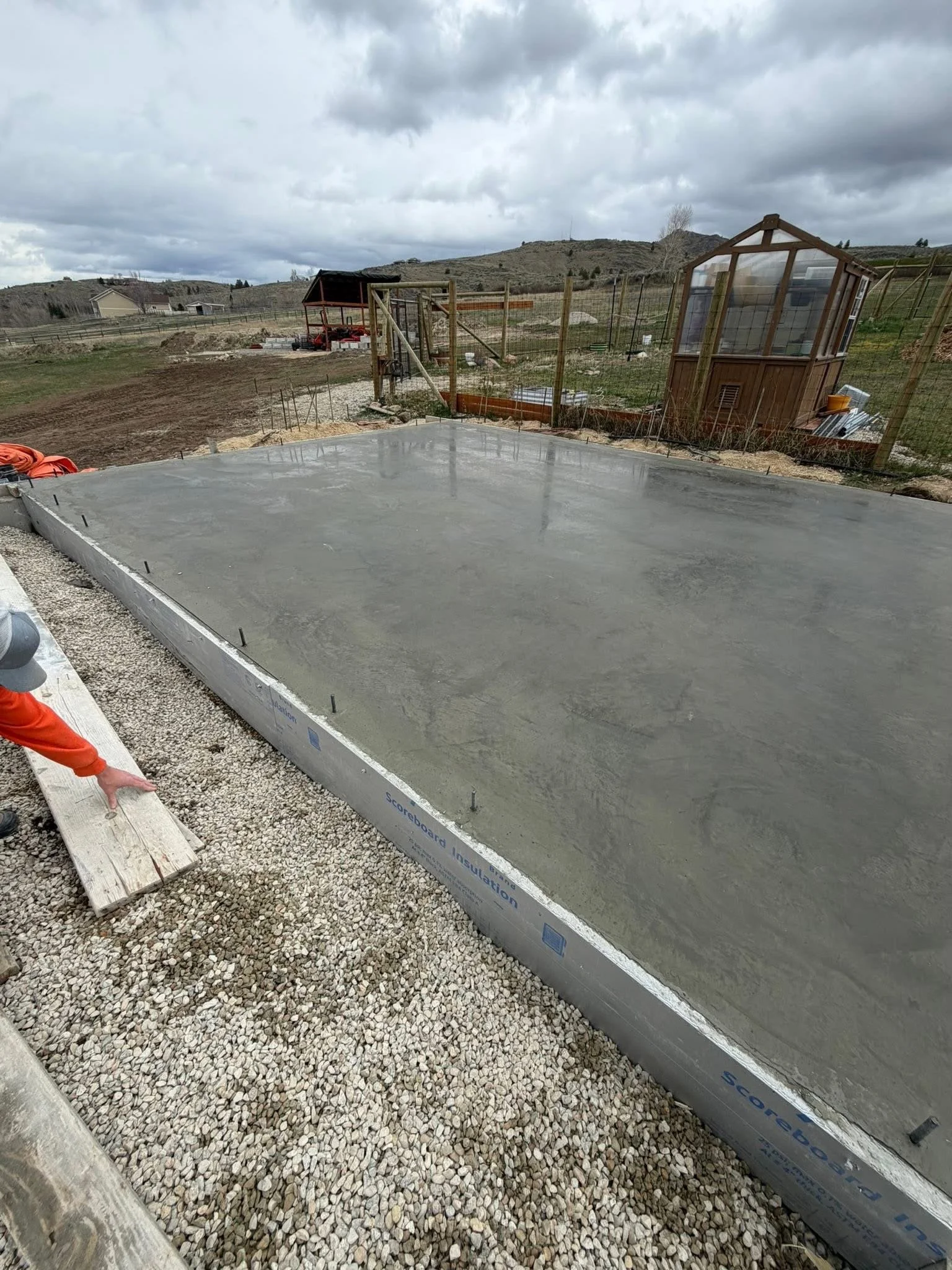 Concrete slab poured on a construction site surrounded by gravel and wooden forms, with hills and cloudy sky in the background.
