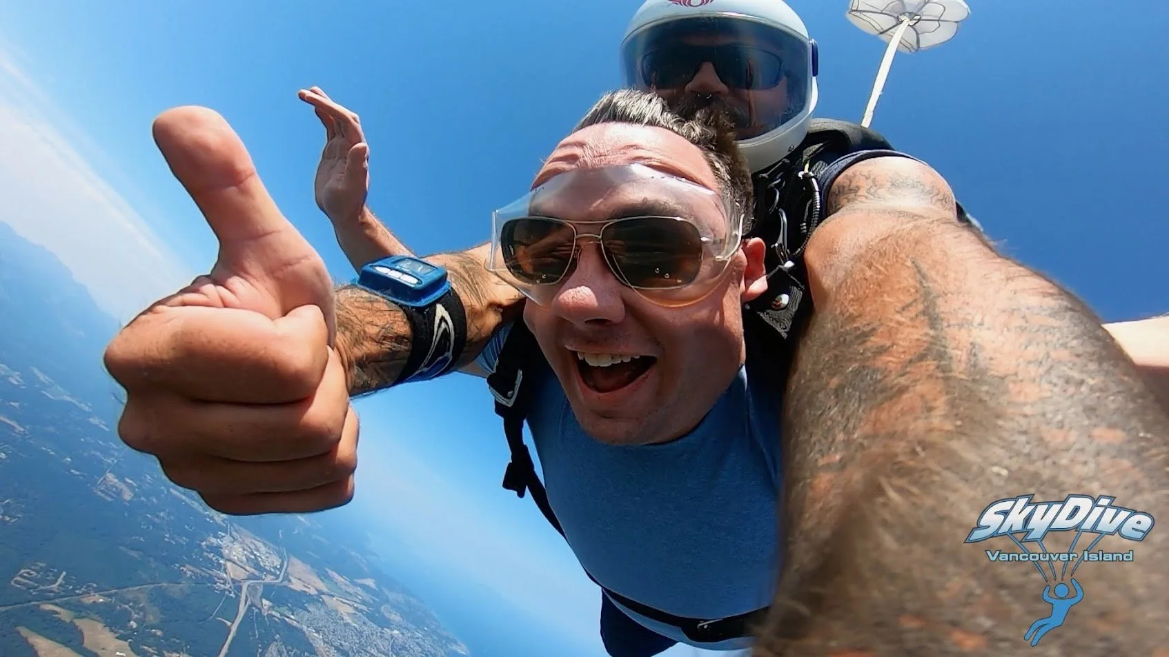 Two skydivers in free fall above the landscape, one giving a thumbs-up, with the Skydive Vancouver Island logo in the corner.