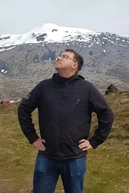 Person standing outdoors on grassy terrain with hands on hips, wearing a black jacket and glasses, with snow-capped mountains in the background.