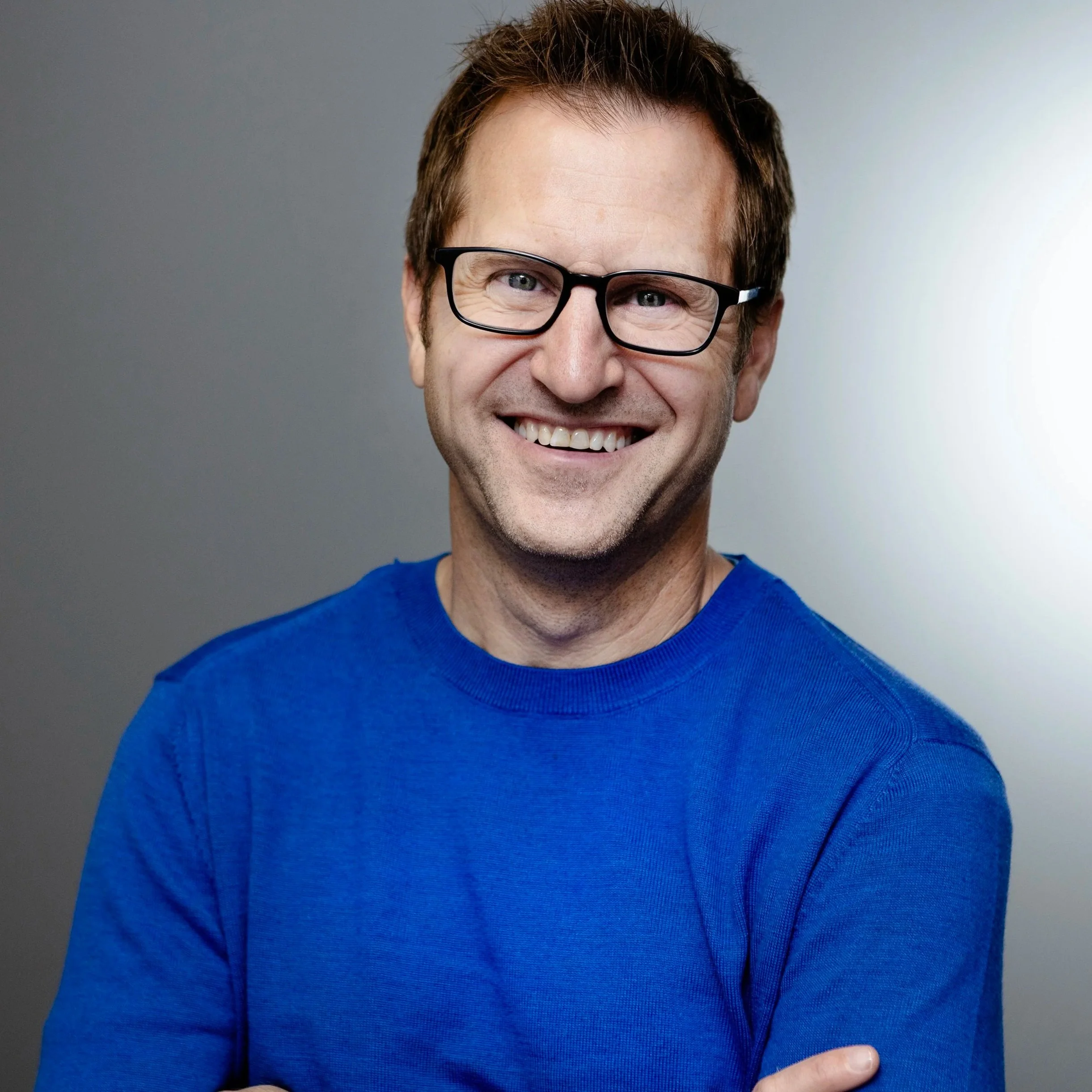 Close-up of a smiling man with brown hair, wearing black glasses and a blue shirt, set against a gray background.
