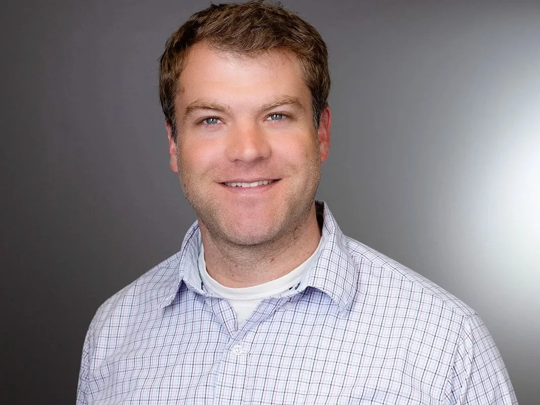 A smiling man with light brown hair wearing a checkered shirt against a gray background.