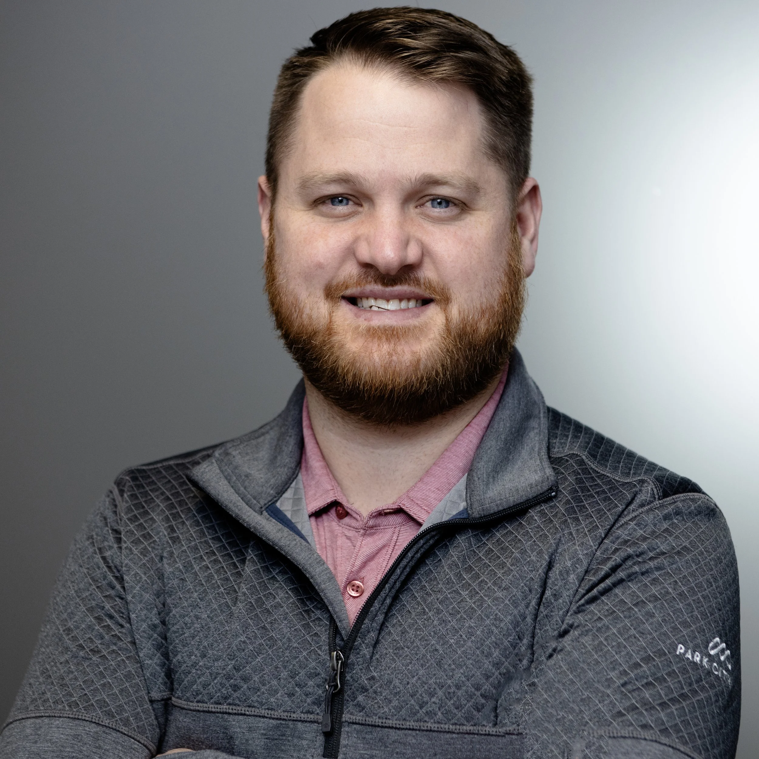 Close-up of a smiling man with blue eyes, red hair, and a beard wearing a gray patterned zip-up jacket over a pink collared shirt. The background is a plain gradient from dark gray to light gray.