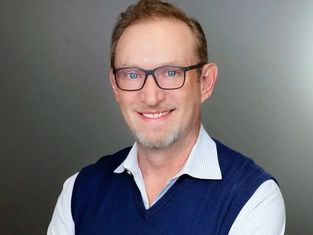 Headshot of a man with glasses, short brown hair, and a goatee, smiling and wearing a white collared shirt with a blue sweater vest against a gray background.