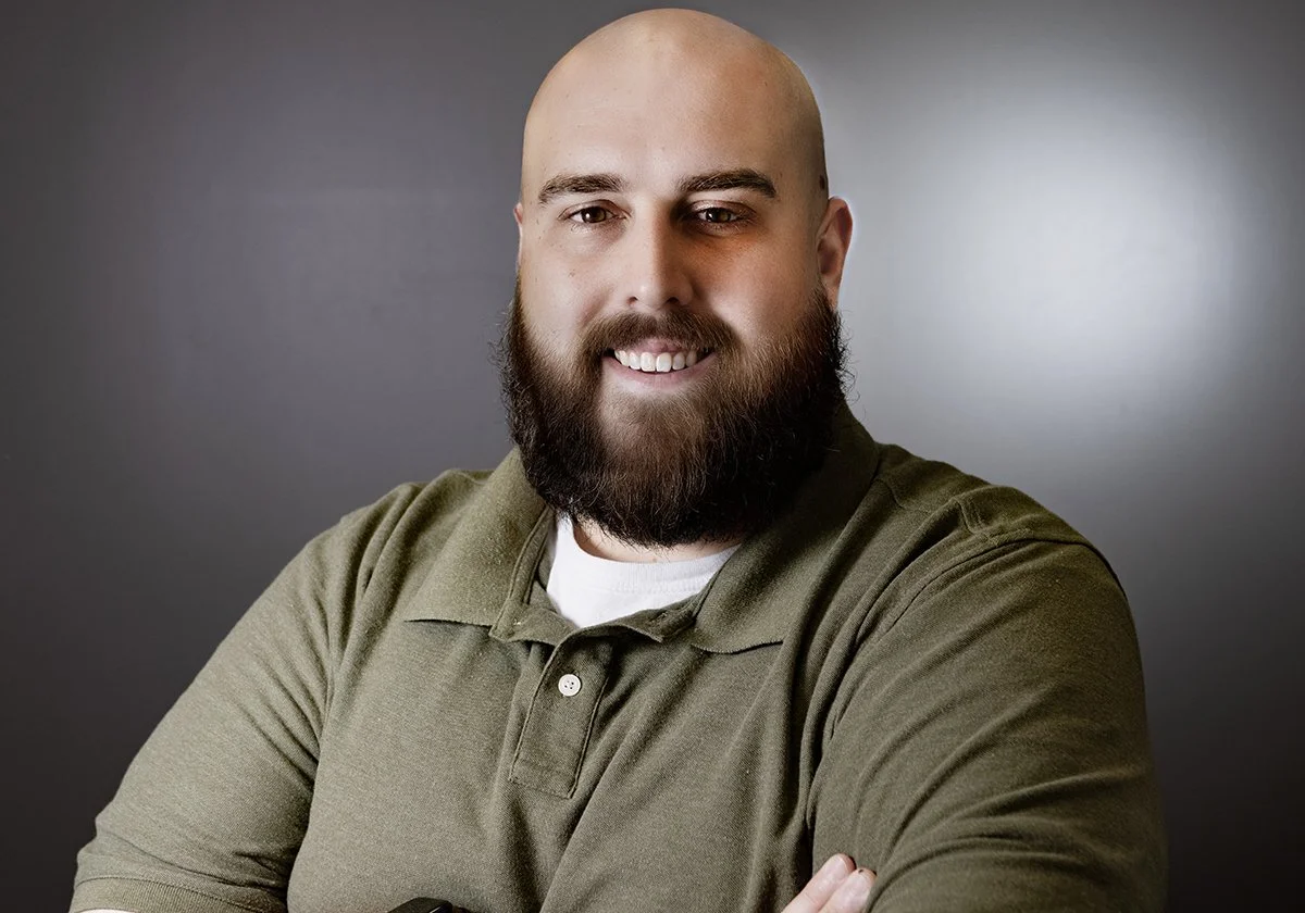 A bald man with a full beard smiling, wearing a green polo shirt, standing against a gray background.