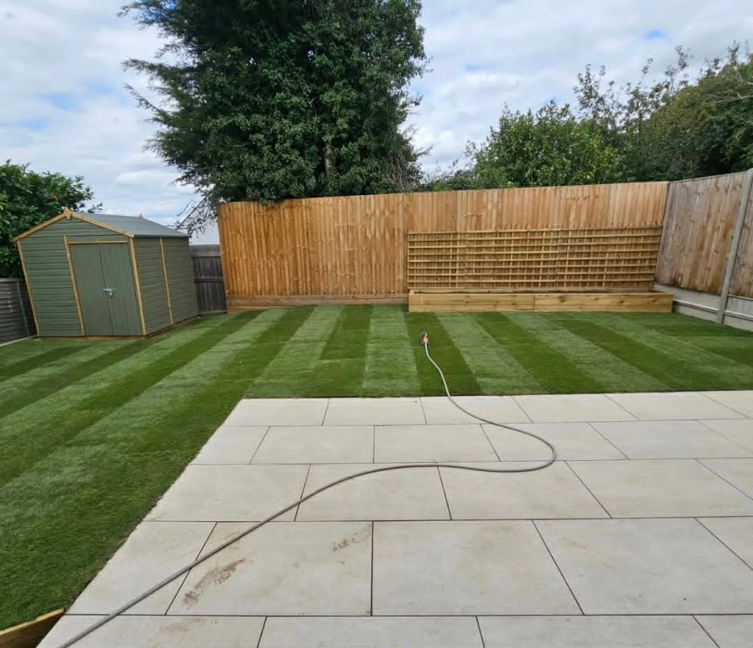 A backyard with freshly laid striped green grass, a concrete patio in the foreground, a wooden shed on the left, and a wooden fence surrounding the yard.