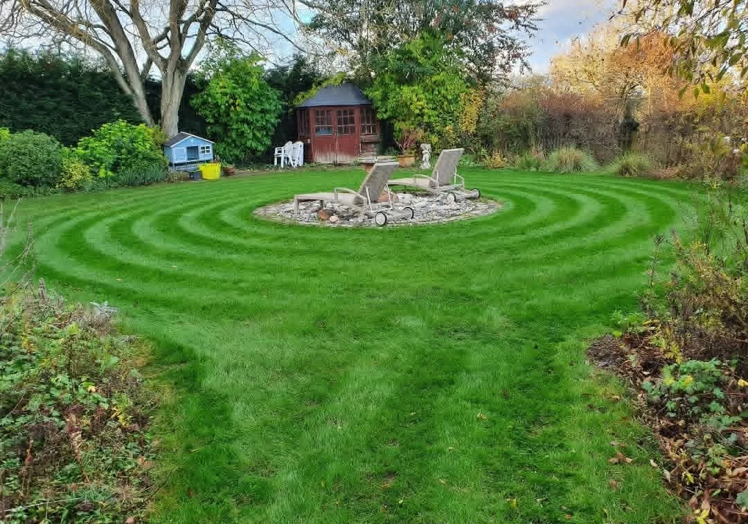 Well-maintained green lawn with curved stripes, centered around a small fire pit with two reclining chairs. A garden shed and trees with fall foliage are in the background.