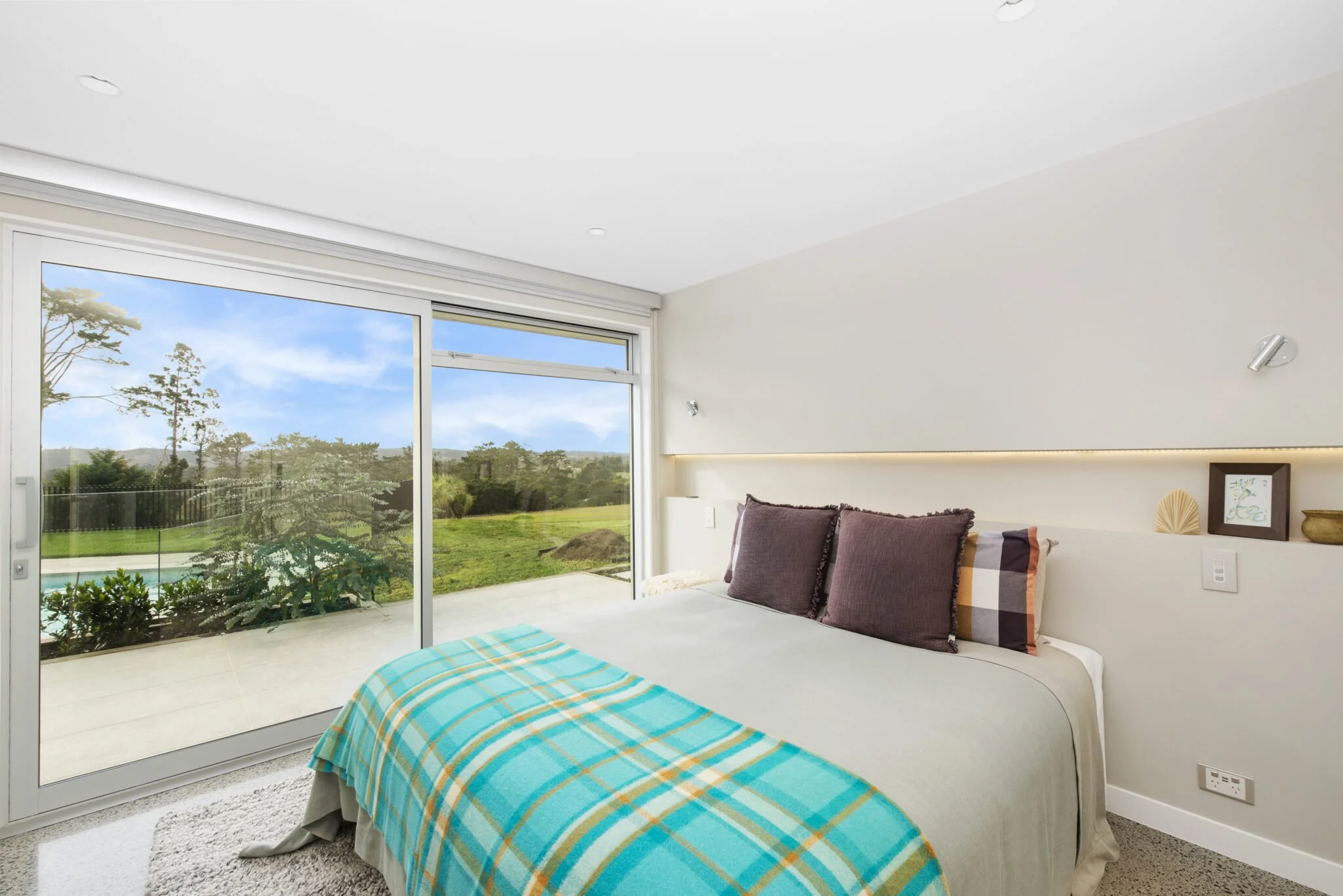 A bedroom with a large sliding glass door that opens to a green outdoor landscape with trees and hills, a bed with a colorful plaid blanket, and purple pillows.