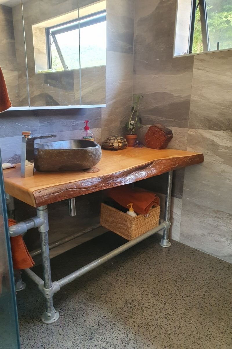 Bathroom with a stone basin sink on a wooden countertop, small decorative plants and objects, mirror, window, and storage basket underneath.