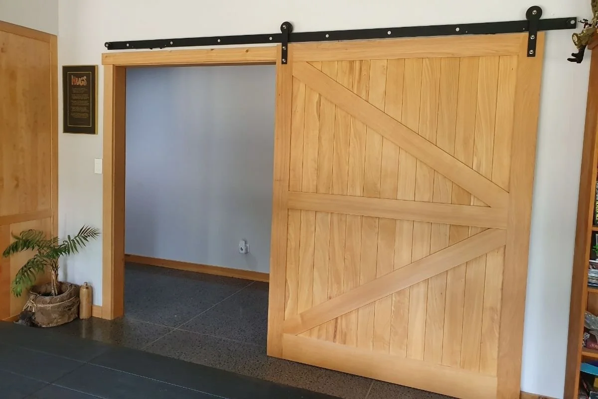 A wooden sliding barn door with a diagonal cross brace, mounted on a black metal rail, partially open to reveal a white wall and electrical outlet inside a room.