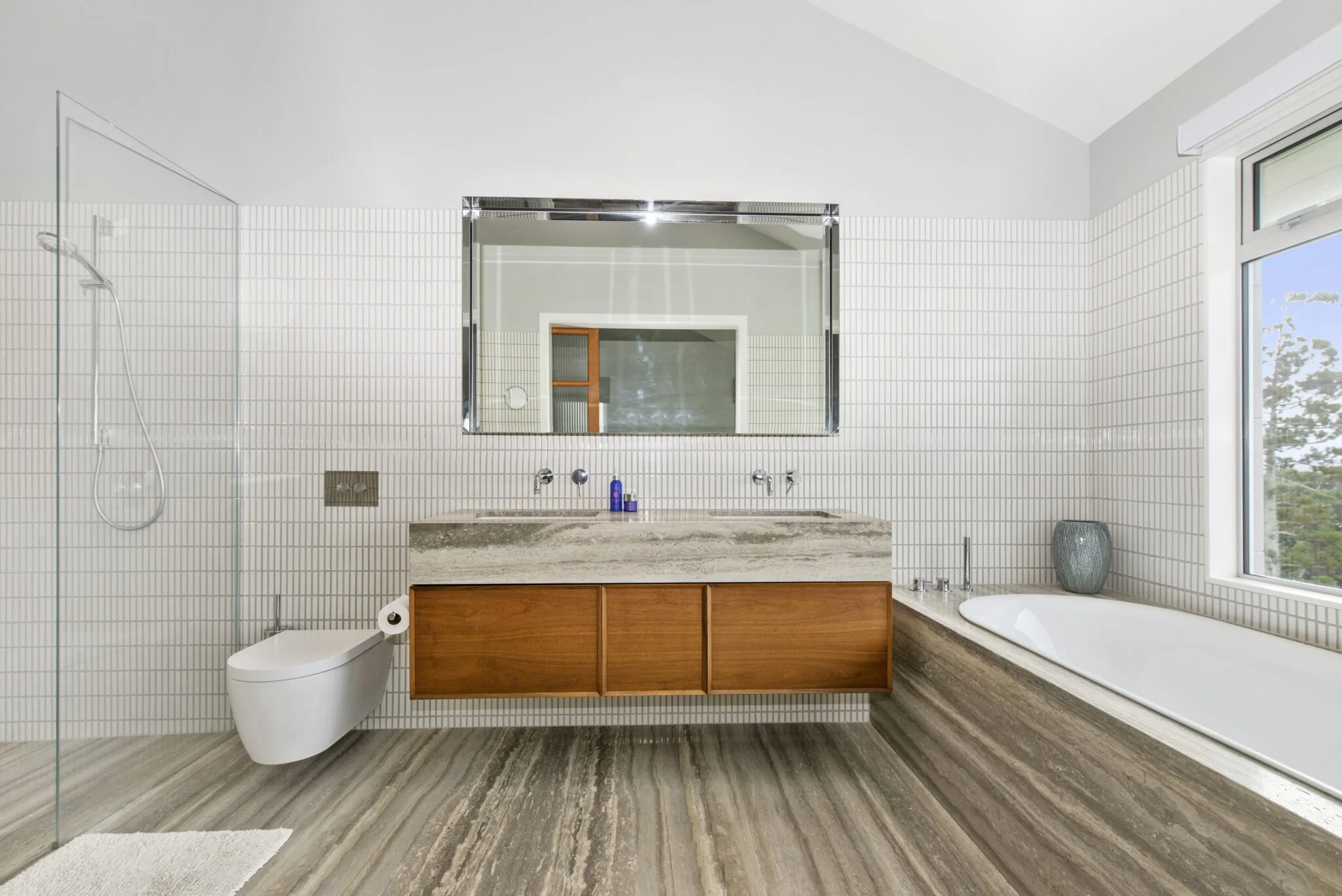 Modern bathroom with white tiled walls, a large mirror above a marble vanity, a toilet, a bathtub near a window, and wooden cabinet beneath the sink.