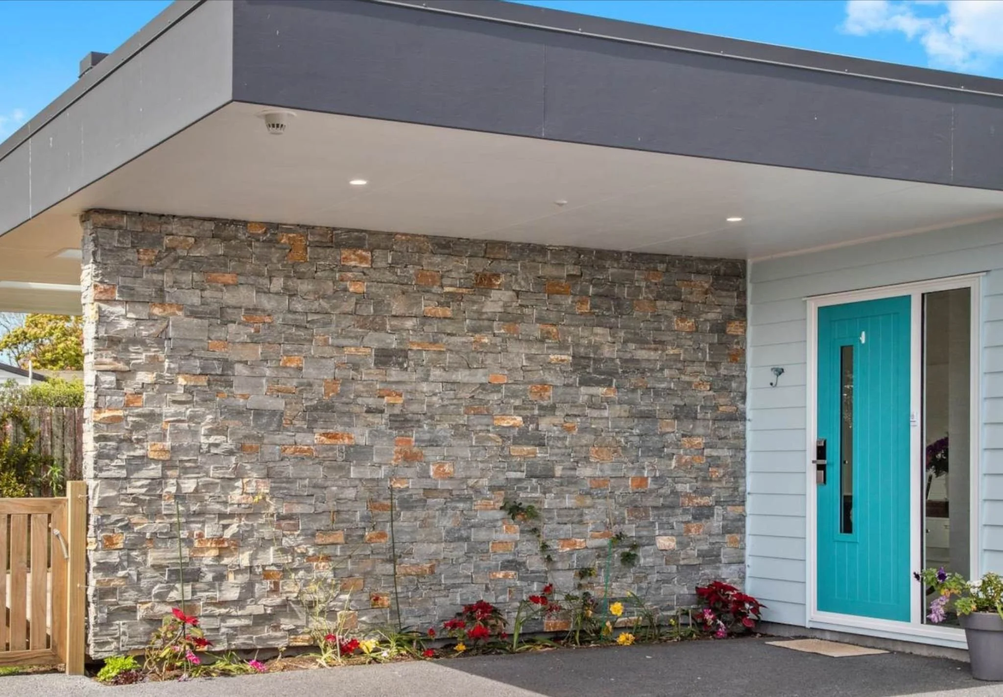 View of modern house exterior with gray stone wall, white addition, and turquoise door, surrounded by colorful flowers.