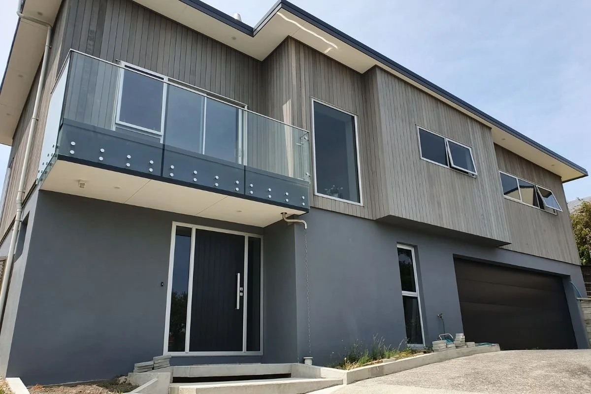 Modern two-story house with a gray exterior, glass balcony, large windows, and a garage. Clear sky overhead.