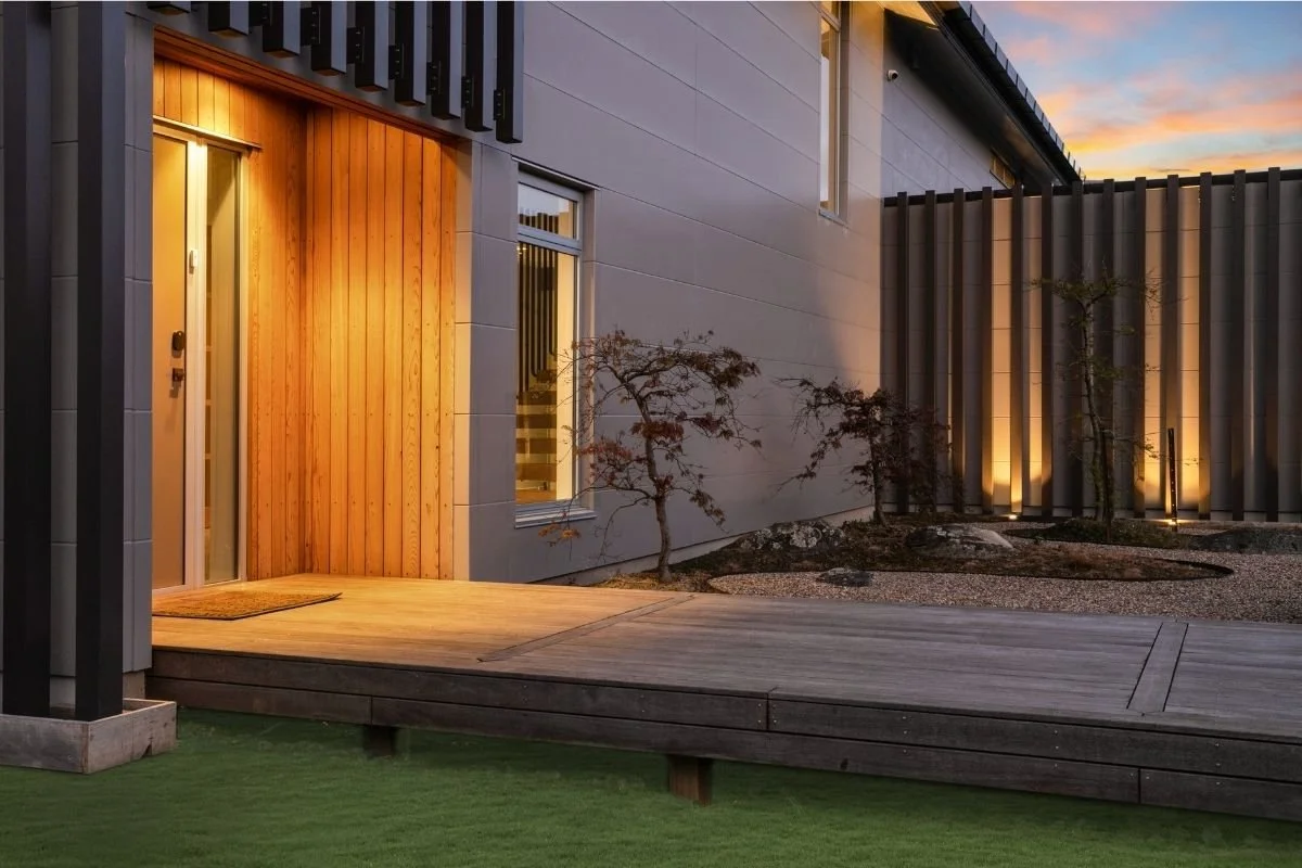 Modern house entrance with a wooden door, a small porch, and minimalist landscaping including small trees and lighting at dusk.