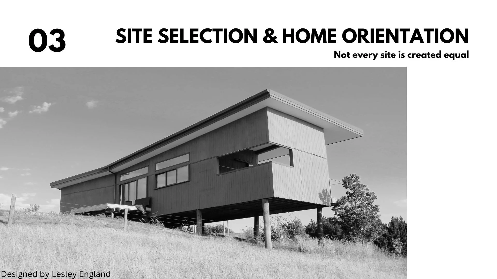 Black and white photo of a modern, elevated house with large windows and flat roof, set on a grassy landscape with trees in the background.
