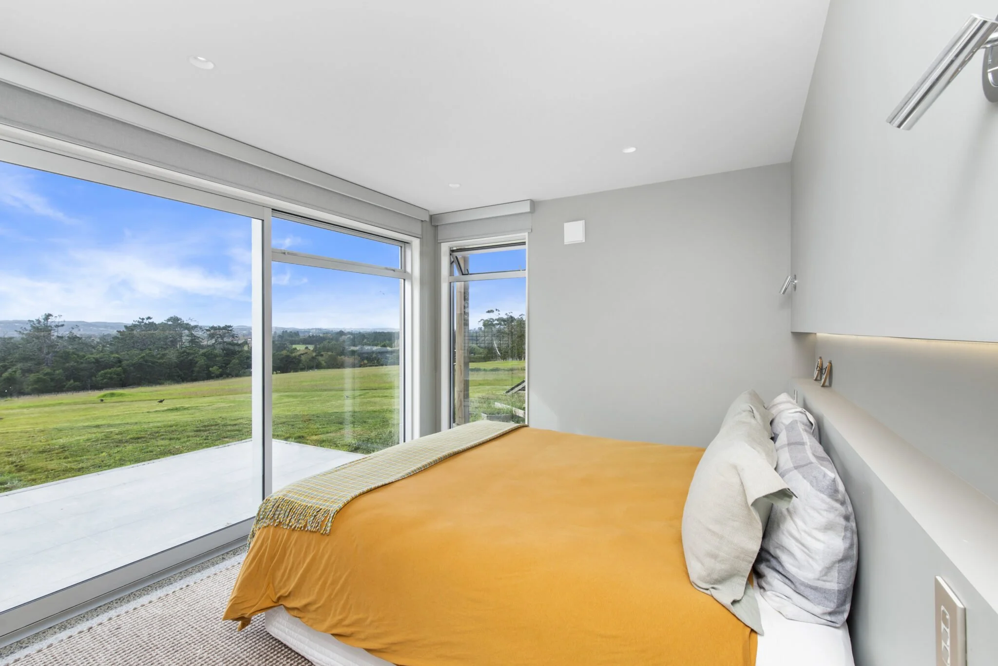 Modern bedroom with large floor-to-ceiling windows overlooking a green landscape and blue sky, featuring an orange bed with checkered pillows and a beige throw blanket.