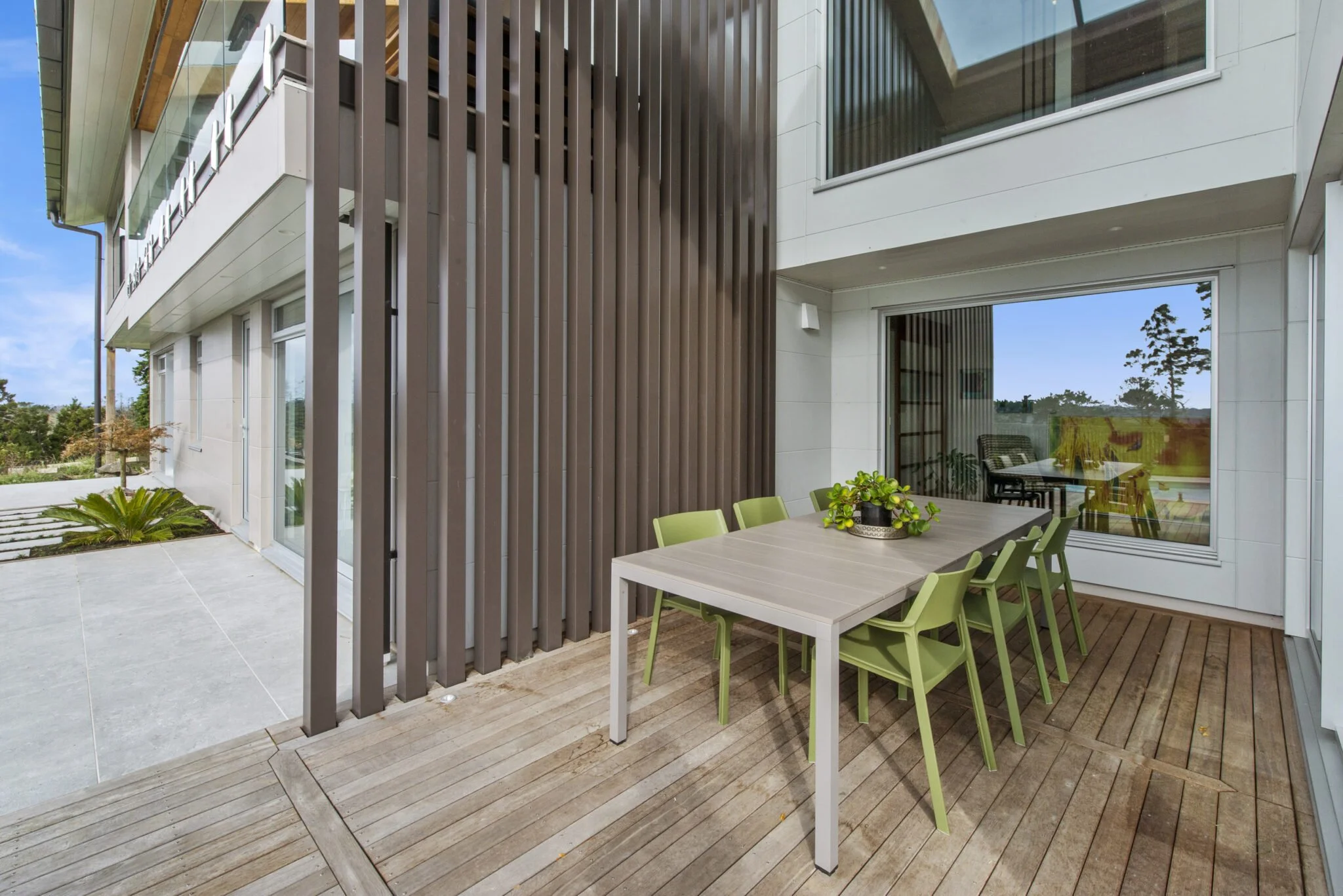 Modern house exterior with a covered patio area, wooden floor, glass sliding door, outdoor dining table with green chairs, and vertical brown wooden slats.