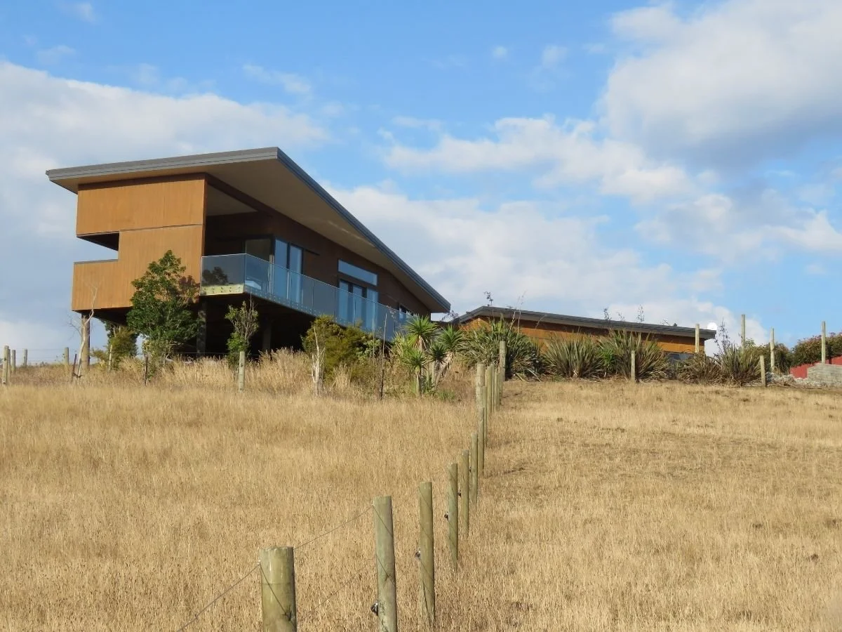 Modern house with wooden exterior, large glass windows and balcony, situated on a grassy hill under a partly cloudy sky.
