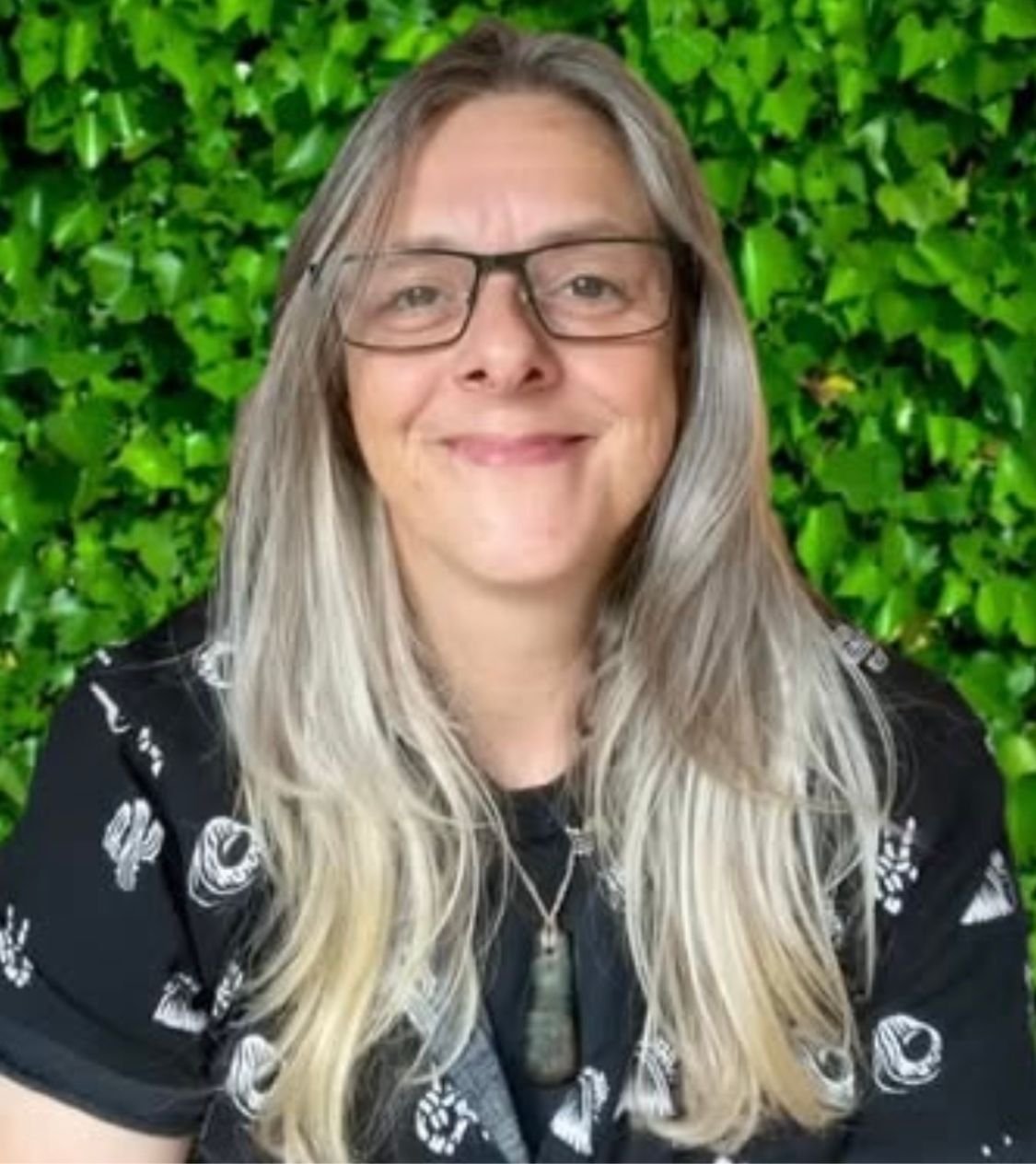 Woman with long blonde hair and glasses smiling in front of a green leafy background.