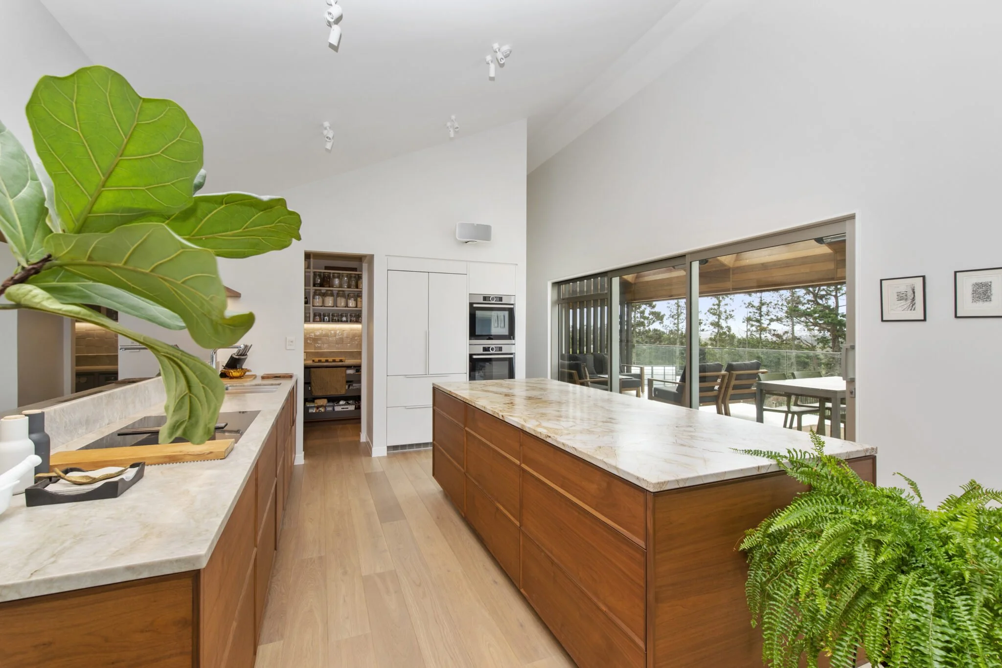 Modern kitchen with white walls, wooden cabinets, a marble kitchen island, large window with outdoor view, and indoor plants.