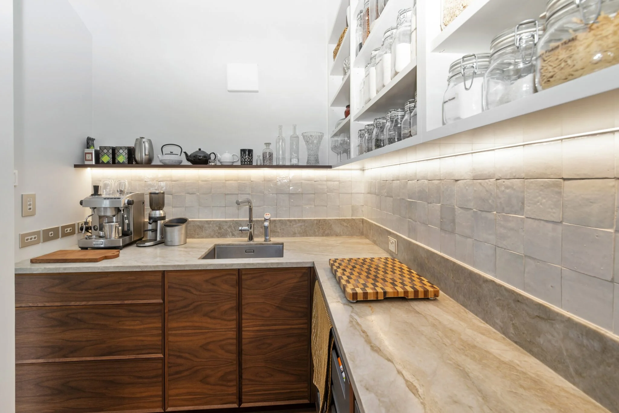 A modern kitchen with white upper cabinets, open shelves filled with glass jars and dishes, a beige marble countertop, a coffee machine, and a small sink.