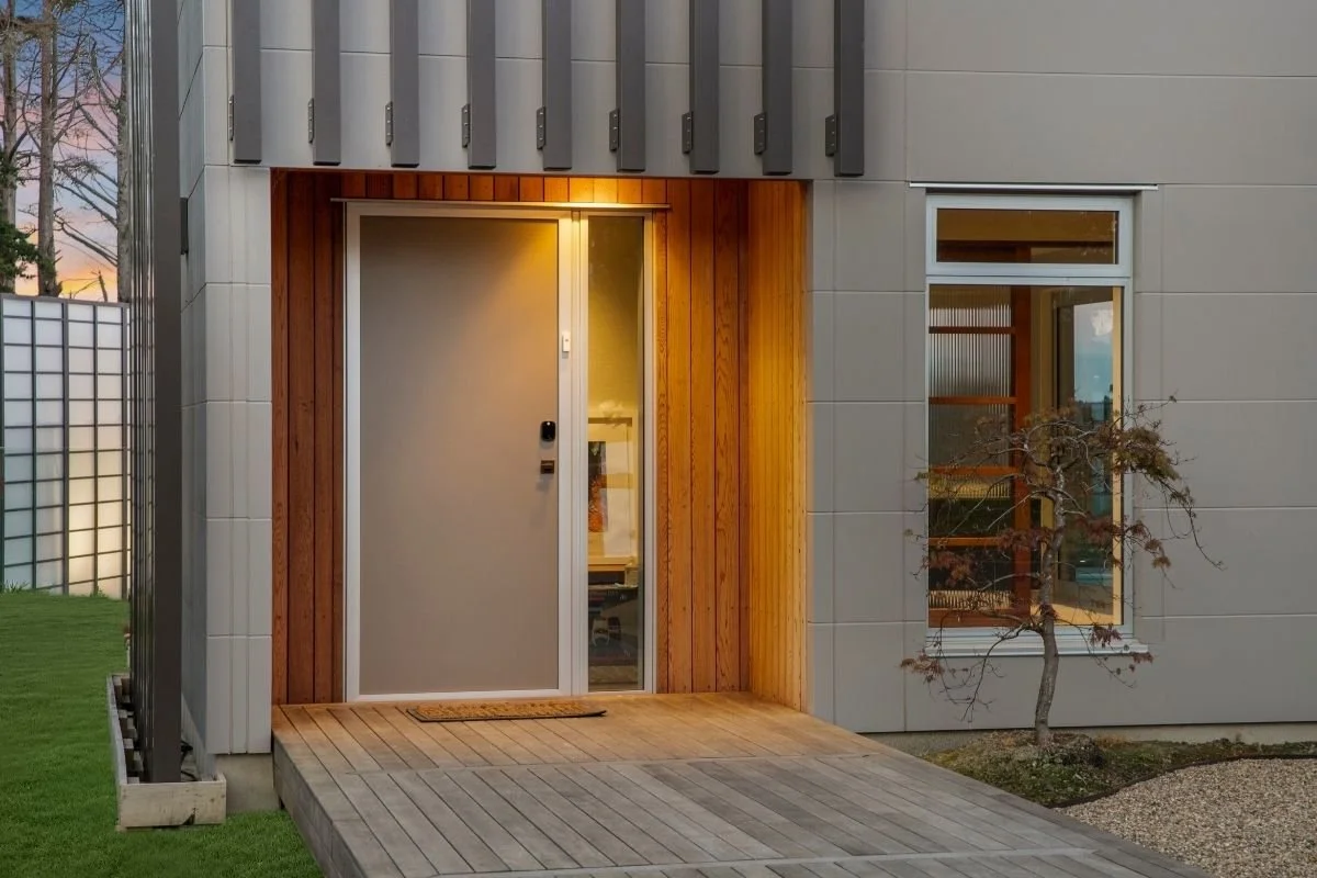 Modern house entrance with a gray door, wooden accents, a small doormat, a window, and a small tree to the right.