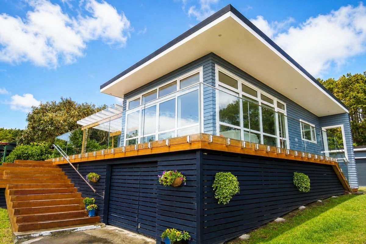 Modern house with large glass windows, a blue exterior, wooden deck, and staircases leading up to the entrance, set against a bright blue sky.
