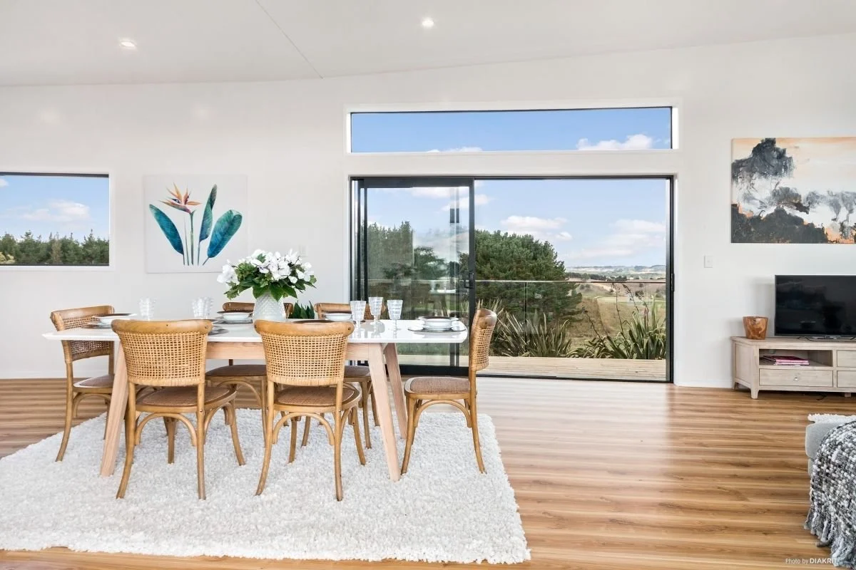 Modern dining room with wooden table, eight wicker chairs, a white vase with white flowers, wooden flooring, large glass sliding door overlooking greenery, and minimalist artwork