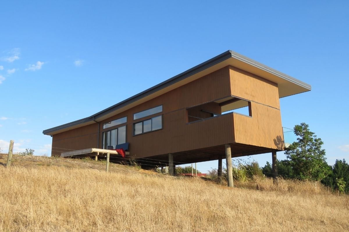 Modern house on stilts with a flat roof, large windows, and wooden exterior, situated on a grassy hill under a clear blue sky.