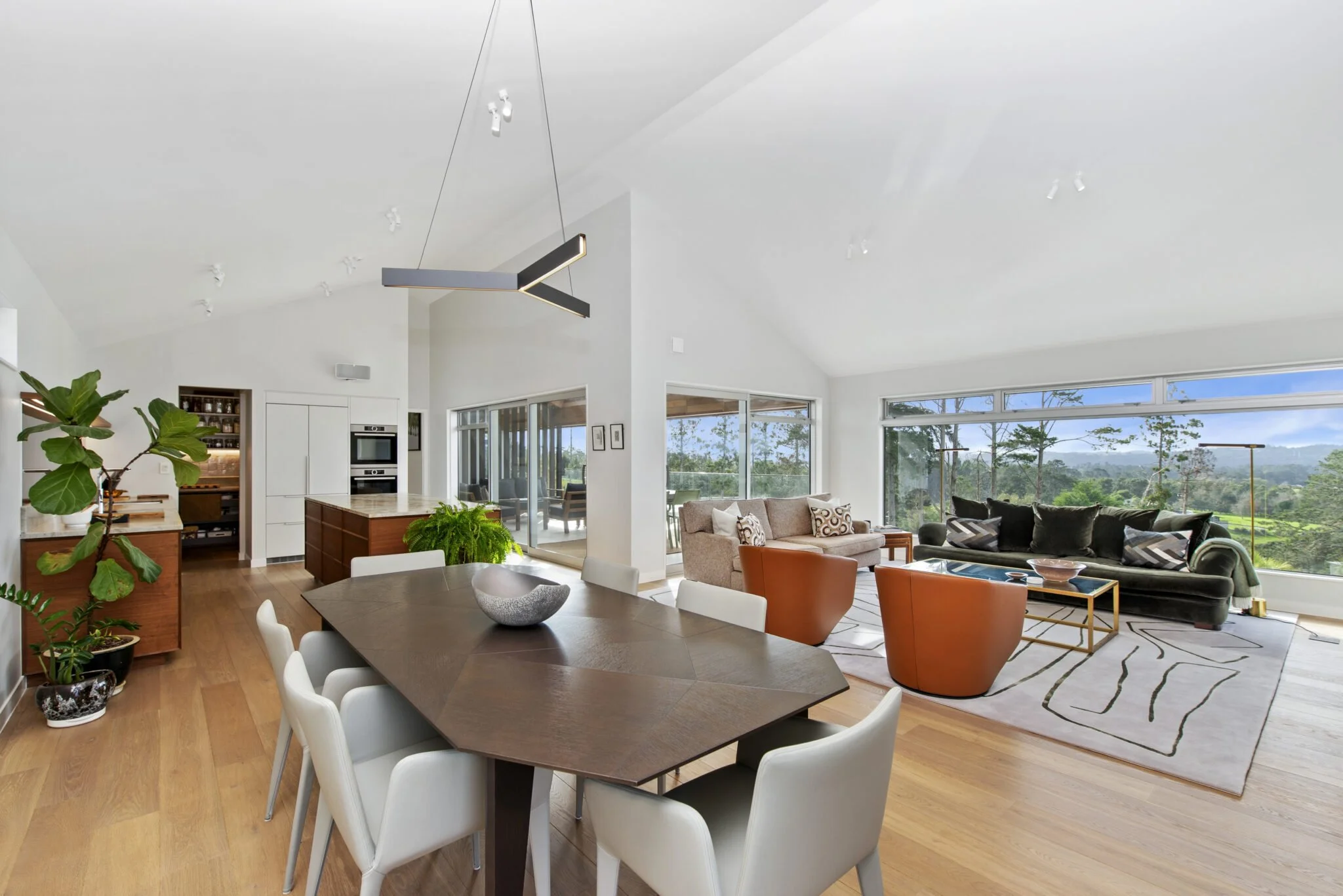 Interior of a bright, modern living room with large windows showing a scenic view, featuring a dining area with a dark wood table and white chairs, a cozy sitting area with a sofa, armchairs, and a rug, and minimalistic decor including plants and pen