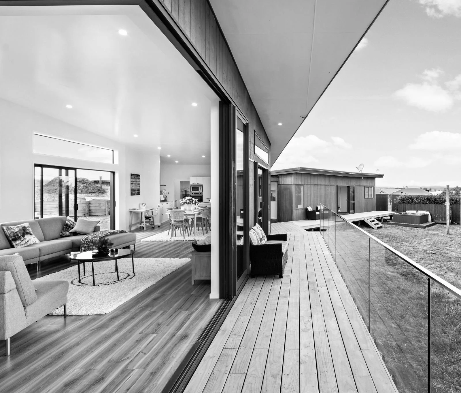 Living room with large glass sliding door opening onto a wooden deck, outdoor hot tub, and backyard. Inside features a sofa, chairs, dining table, and kitchen.