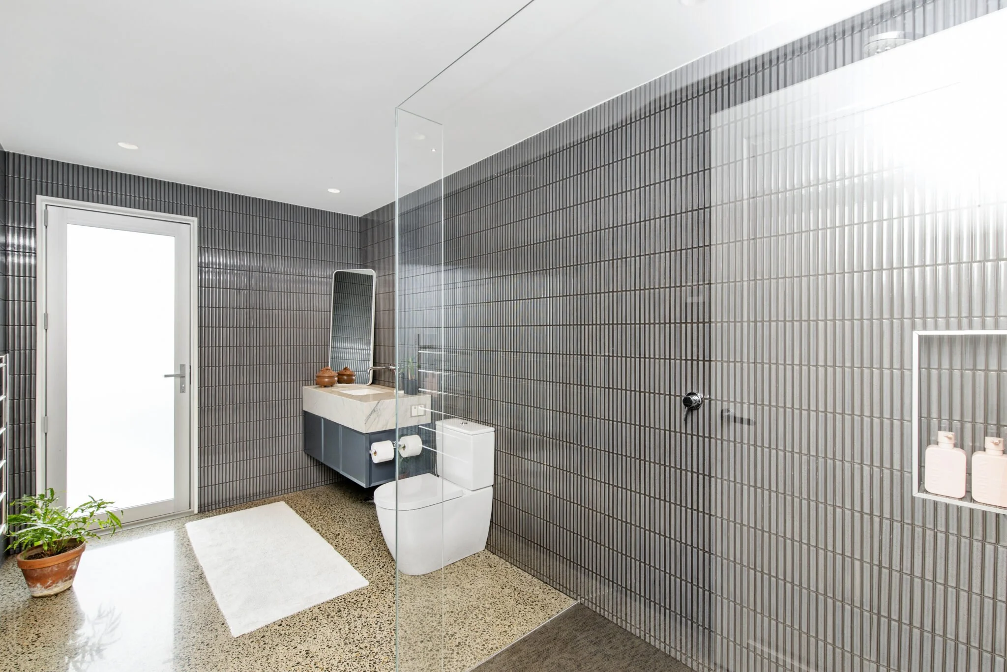 Modern bathroom with black tiled walls, white toilet, marble vanity with mirror, and potted plant near the door.