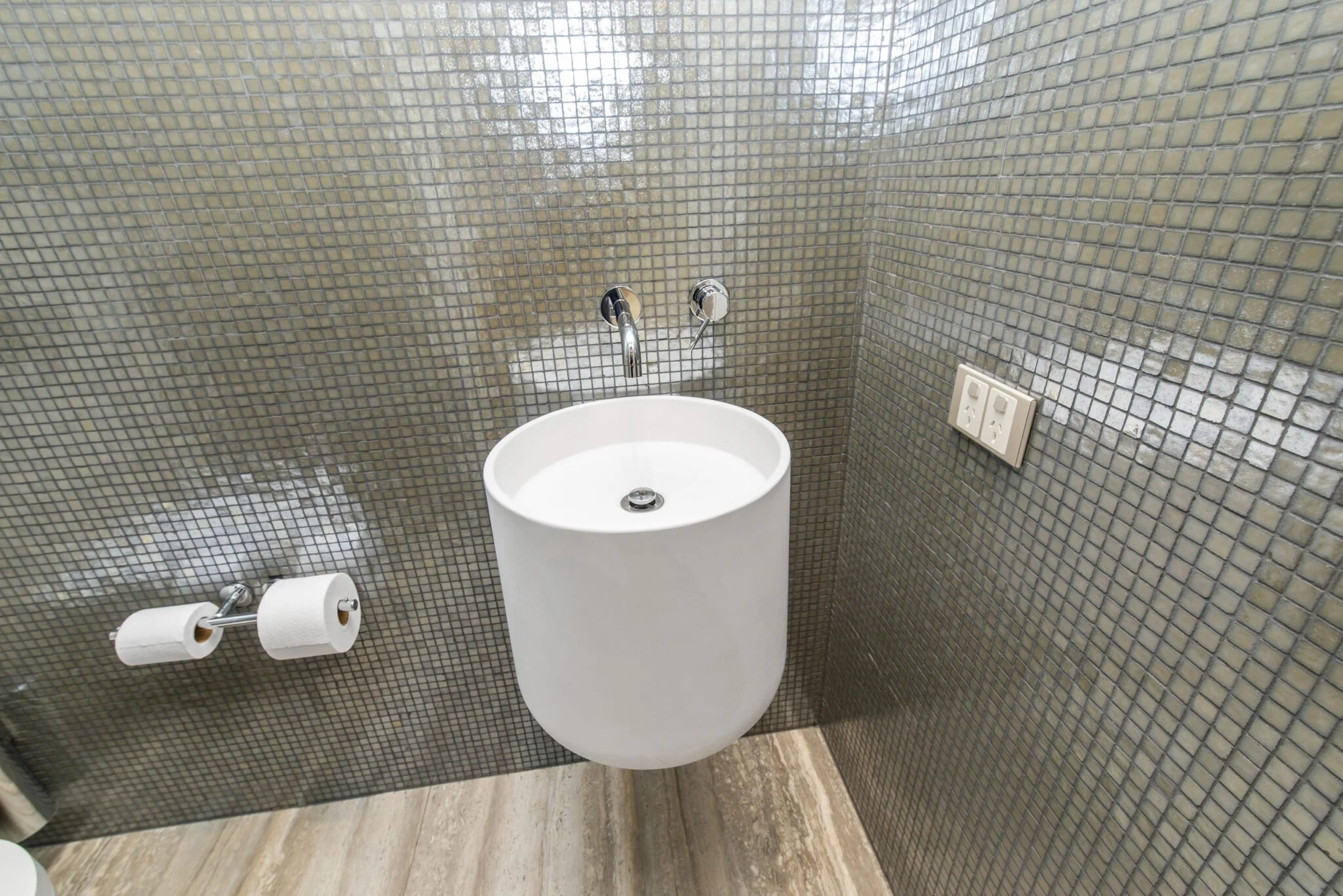 A bathroom wall with small, square, reflective mosaic tiles in shades of brown and beige, with a white toilet and toilet paper holder below, and two power outlets on the right side.
