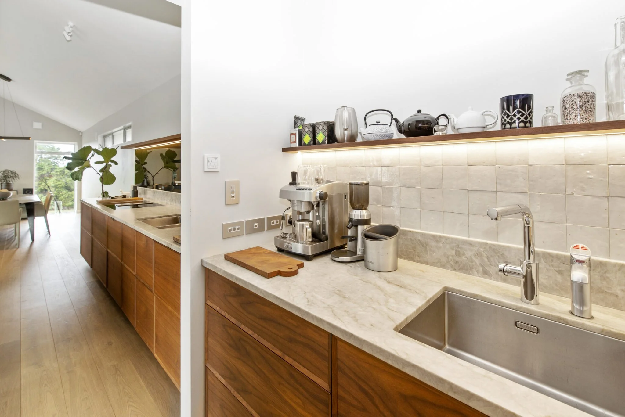 View of a modern kitchen with wooden cabinets, a white marble countertop, coffee-making appliances, and a shelf with decorative jars and teapots, overlooking a dining area with plants and large windows.
