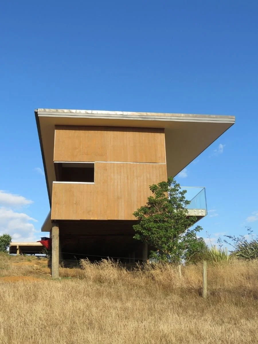 Modern house with a distinctive angular roof, wooden facade, and balcony with glass railing, situated on a grassy hillside under a clear blue sky.