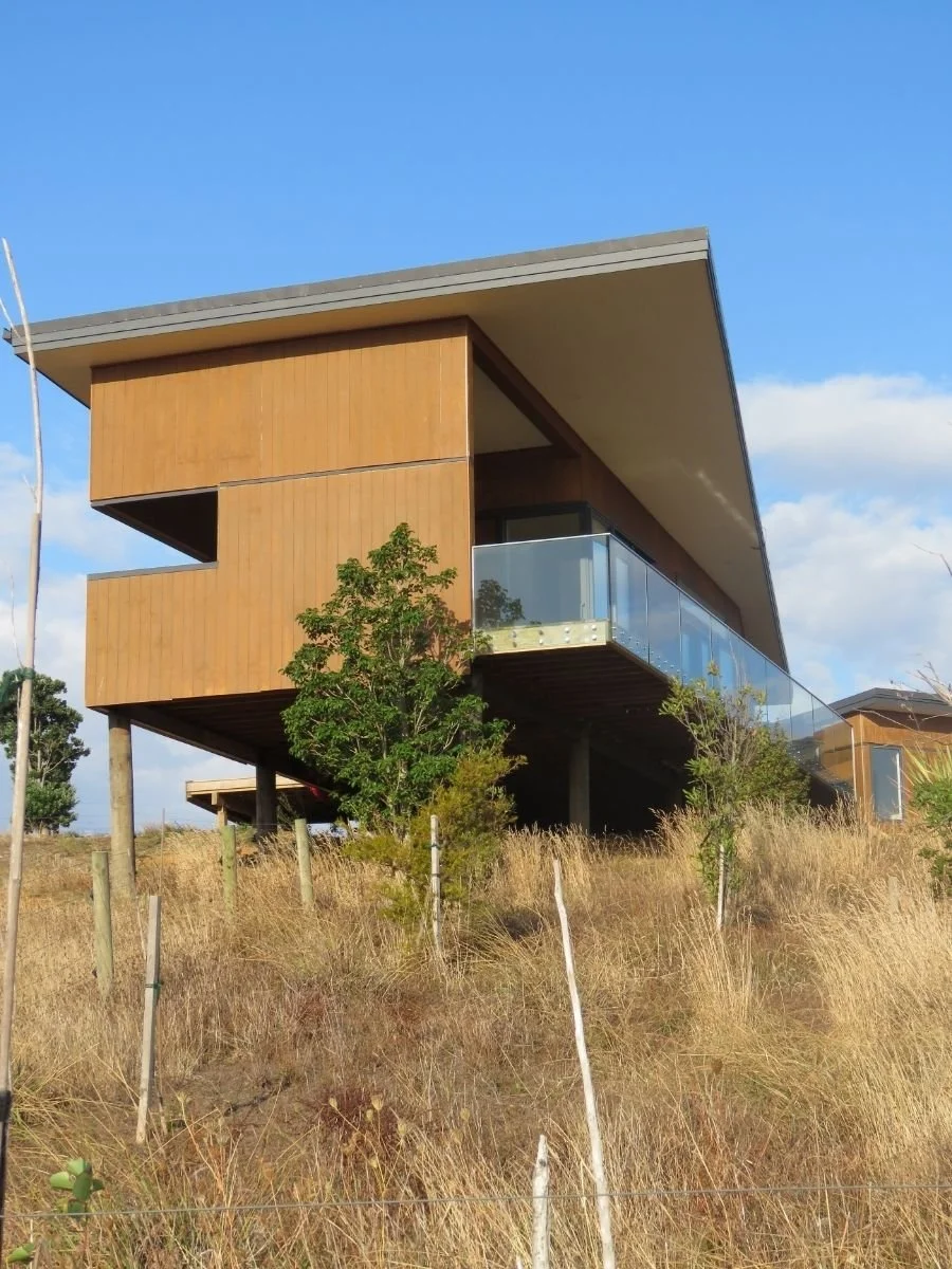 Modern house on stilts with wooden exterior, glass balcony, and slanted roof, surrounded by dry grass and sparse trees under a blue sky.