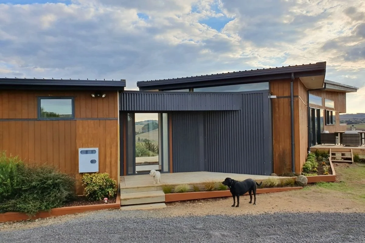 Modern house with wooden and dark gray siding, small front porch, two dogs standing outside, and a landscaped garden with gravel and stepping stones.
