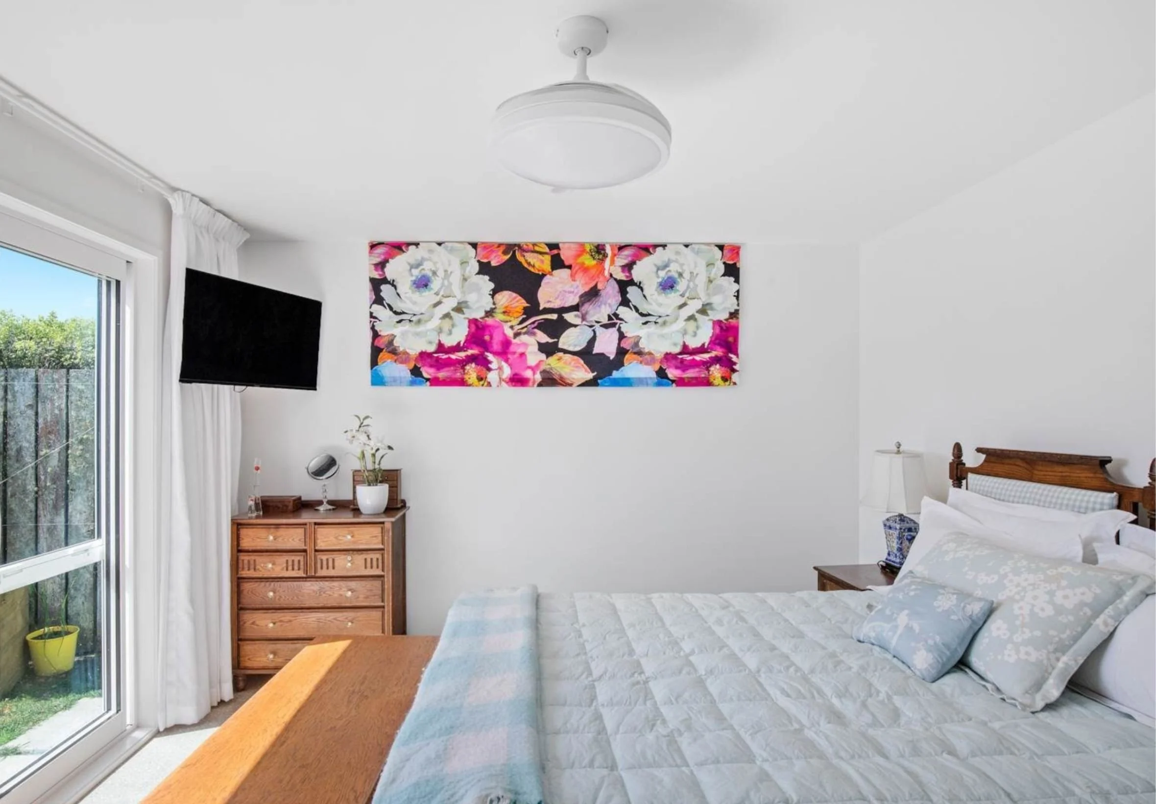 A bedroom with a bed, wooden dresser, and a colorful floral wall art piece above the dresser. There is a sliding glass door with white curtains, letting in natural light. A TV is mounted on the wall next to the door. The bed has a white patterned bed