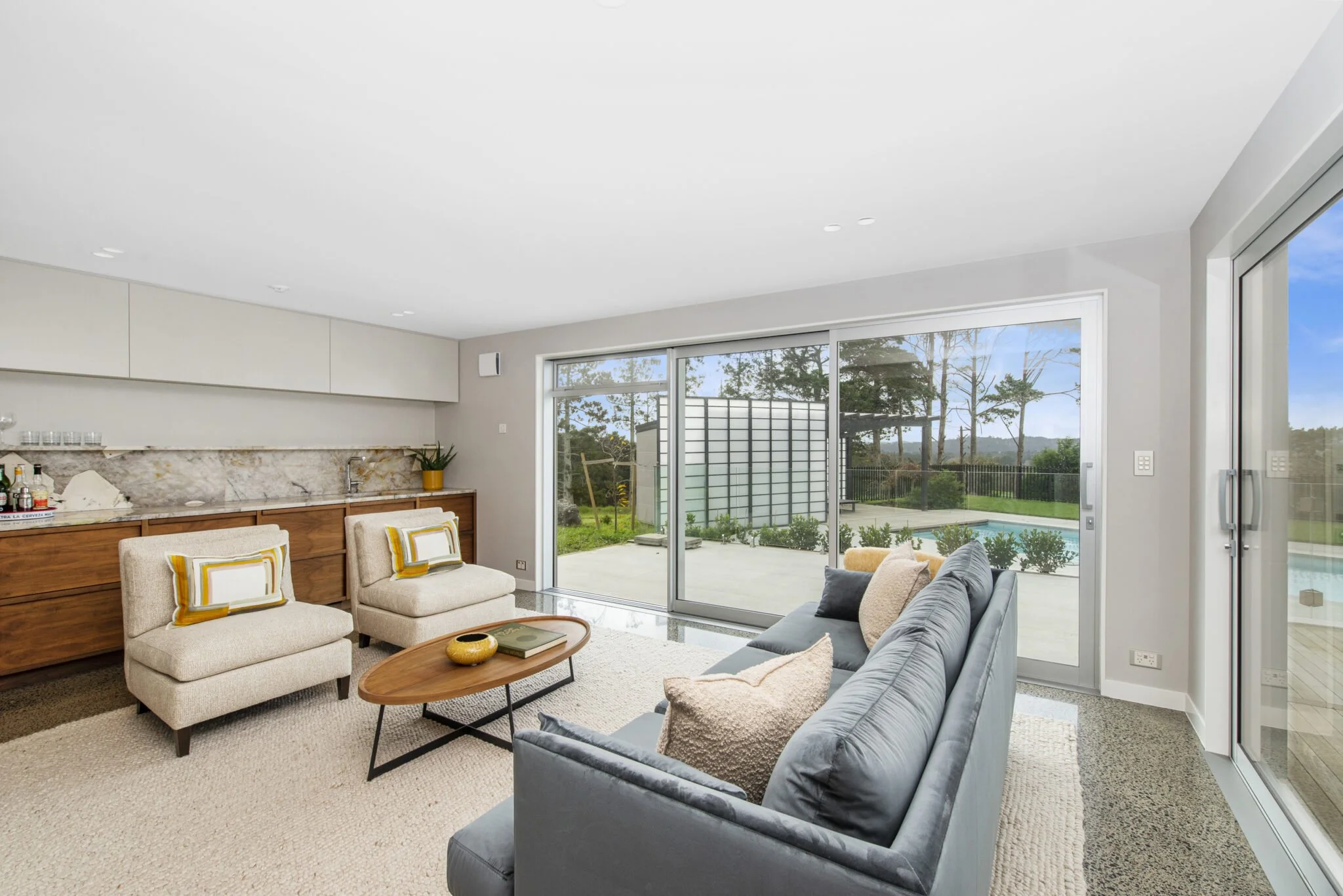 Living room with beige armchairs, gray sofa, wooden coffee table, and sliding glass doors leading to a backyard with a swimming pool, outdoor patio, and trees.