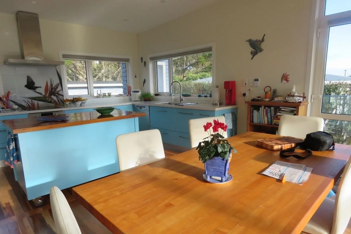 A cozy kitchen and dining area with a wooden table, white chairs, bright blue cabinets, and a window overlooking trees. The table has a potted plant with red flowers, and there are various kitchen appliances and books around.