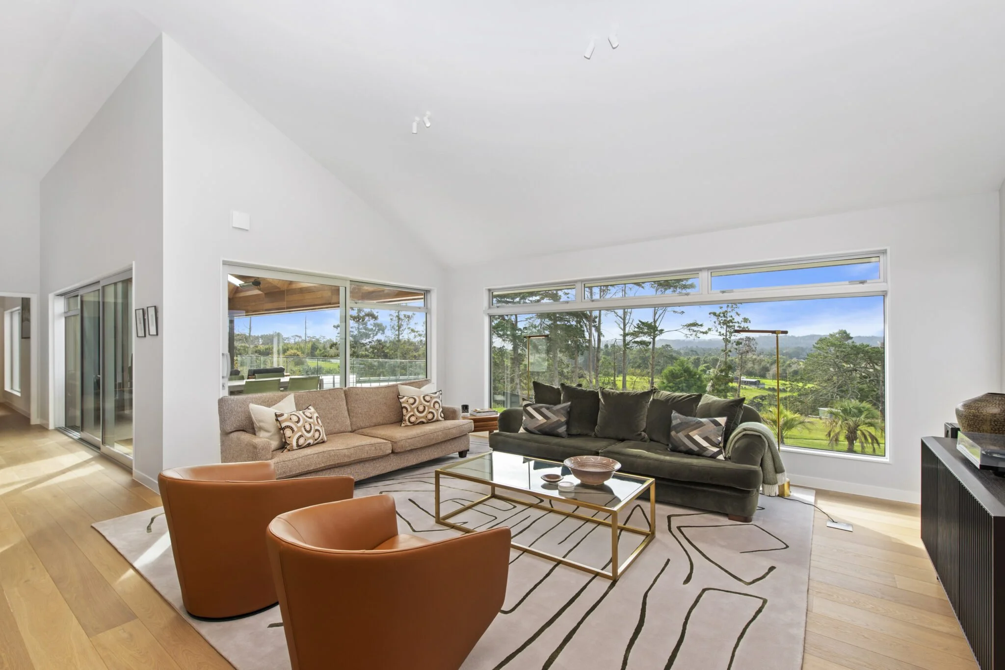 Open living room with large windows, a beige sofa with patterned pillows, black sofa, orange chairs, glass-top coffee table with decorative items, and a view of greenery outside.
