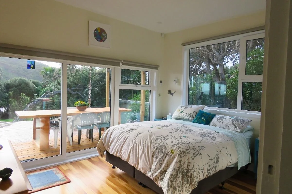 Bedroom with large windows showing outdoor greenery, a bed with floral bedding, and a balcony with a wooden table and white chairs.
