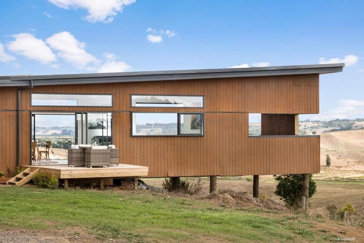 Modern wooden house elevated on stilts with a deck, large windows, and a view of rolling hills in the background under a partly cloudy sky.