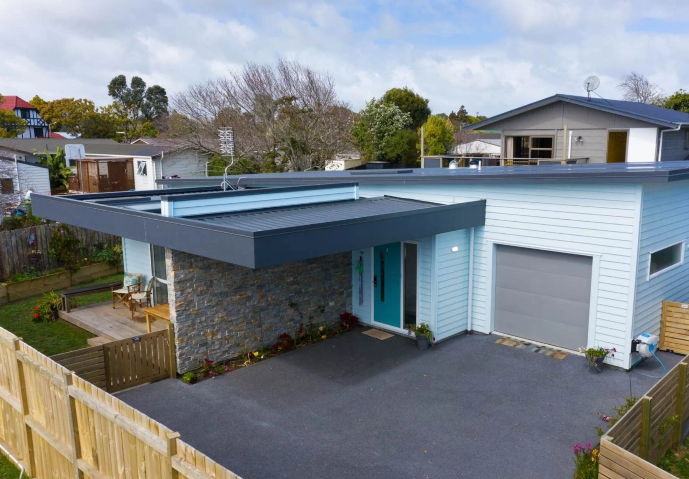 A modern house with a grey roof and light blue siding, a fenced backyard with a patio, a stone accent wall, and a small garden area. Trees and neighboring houses are visible in the background under a partly cloudy sky.