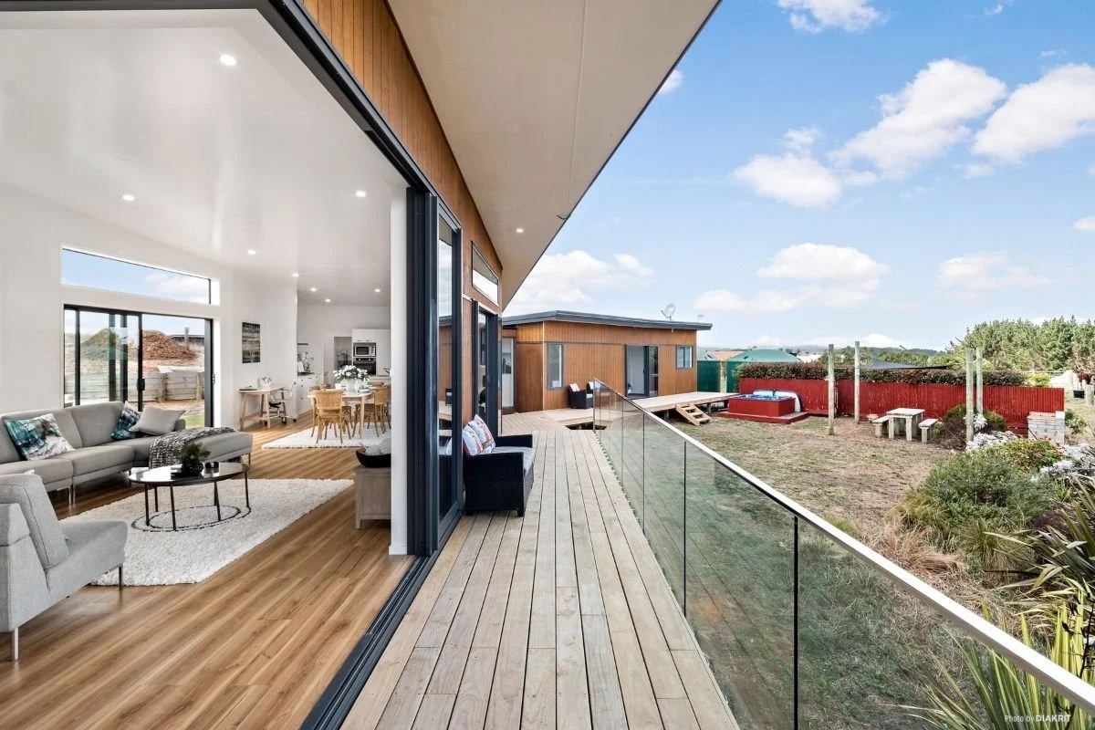 View of a modern interior living room and dining area with large sliding glass doors opened to a spacious wooden deck outside, overlooking a backyard with a hot tub and garden area under a blue sky with scattered clouds.