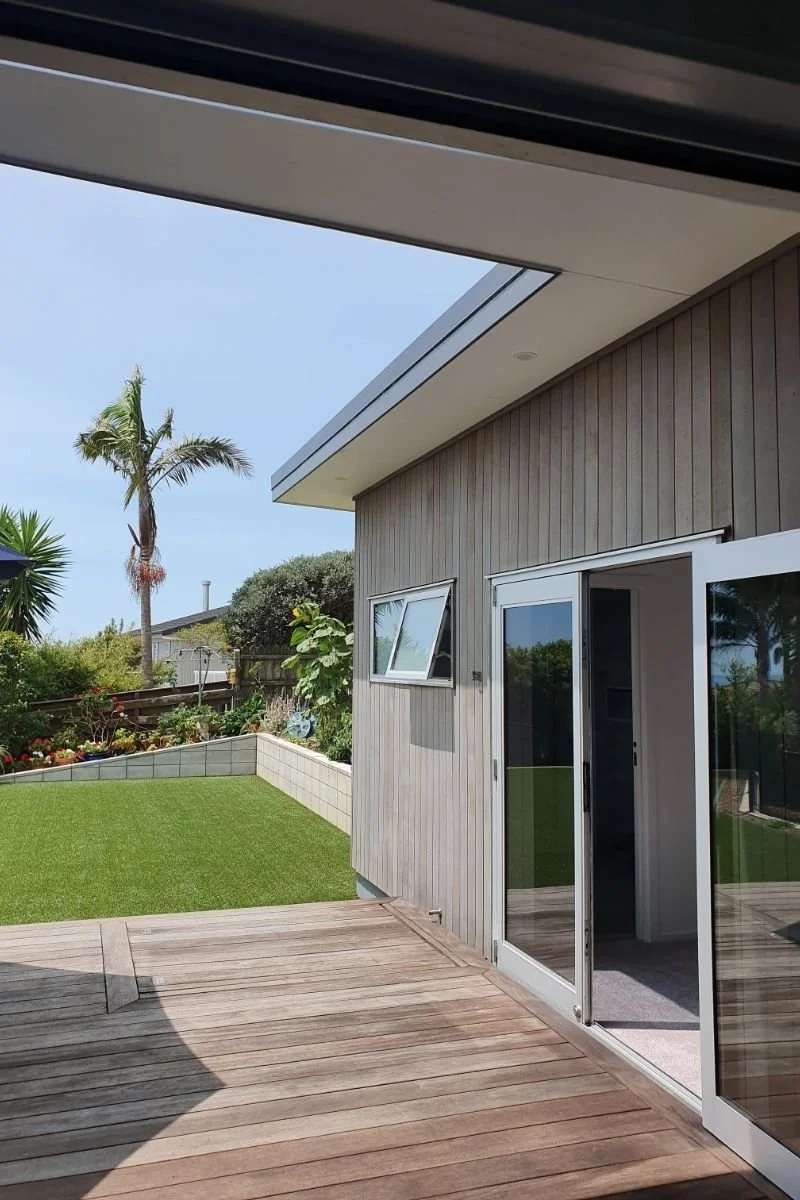 View of a backyard with a wooden deck, sliding glass door, small grassy area, and tropical plants including a palm tree.