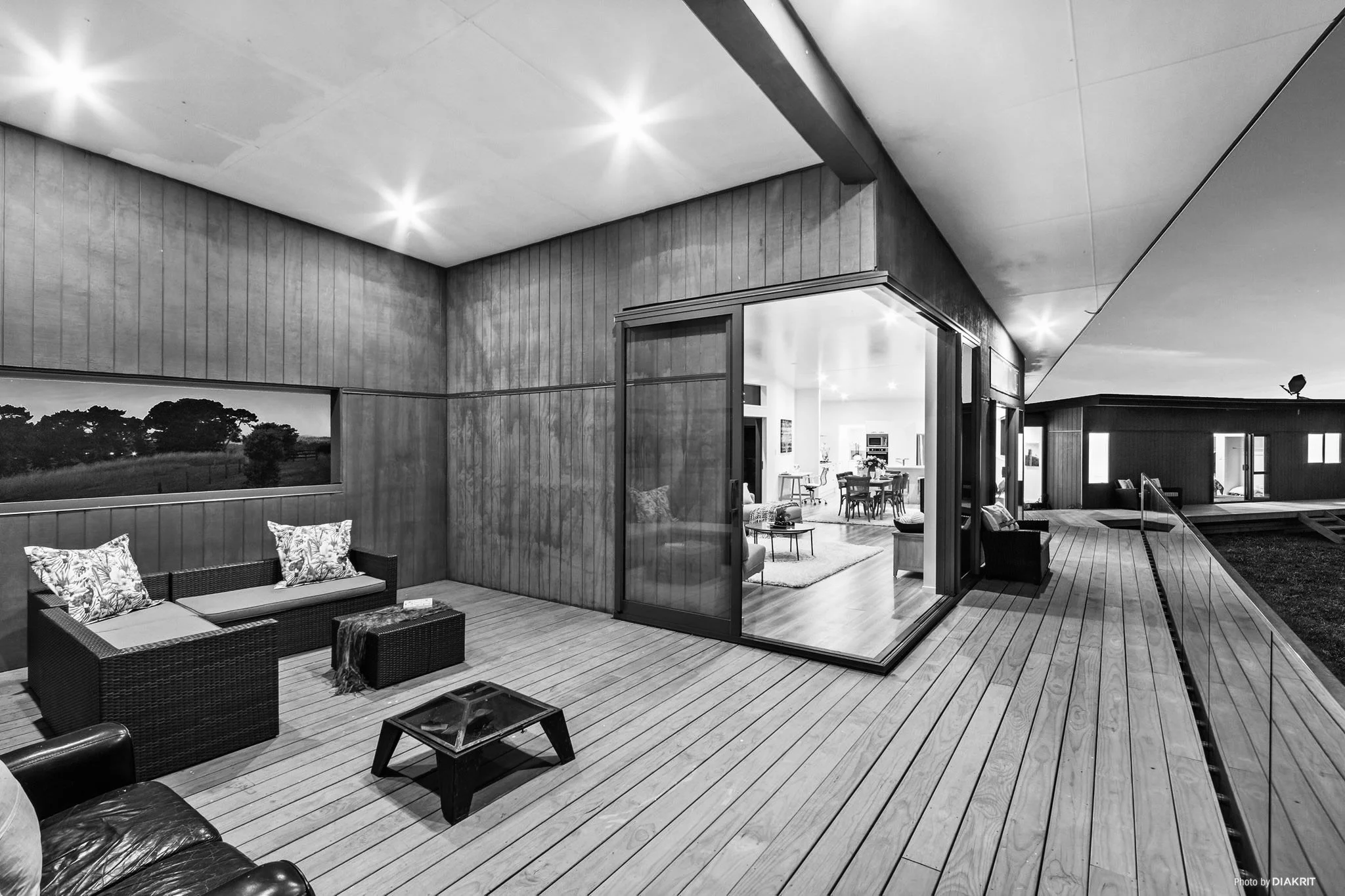 Black and white photo of a modern house interior with a wooden deck patio visible through large glass sliding doors. The patio is furnished with outdoor seating, including a wicker sofa with patterned cushions, simple chairs, and low tables. Inside, there is a cozy living area with a dining table, chairs, and a kitchen in the background. The ceiling has mounted lights, and the overall design is contemporary and open.