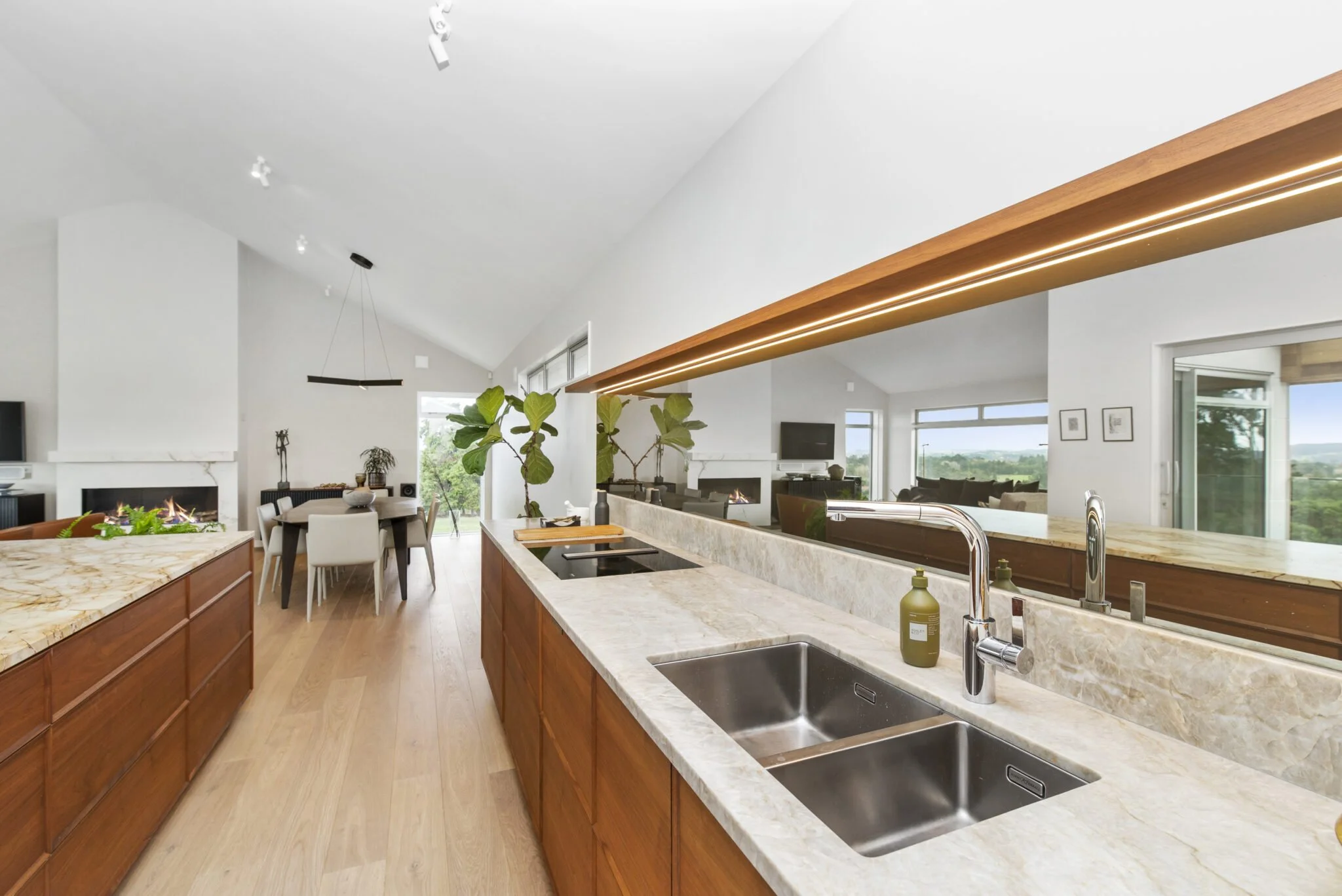 Open concept kitchen with light-colored marble countertops, wooden cabinets, and stainless steel sink. There is a large island with a marble top, modern lighting fixtures, and a view of the living area with a fireplace and large windows showing green