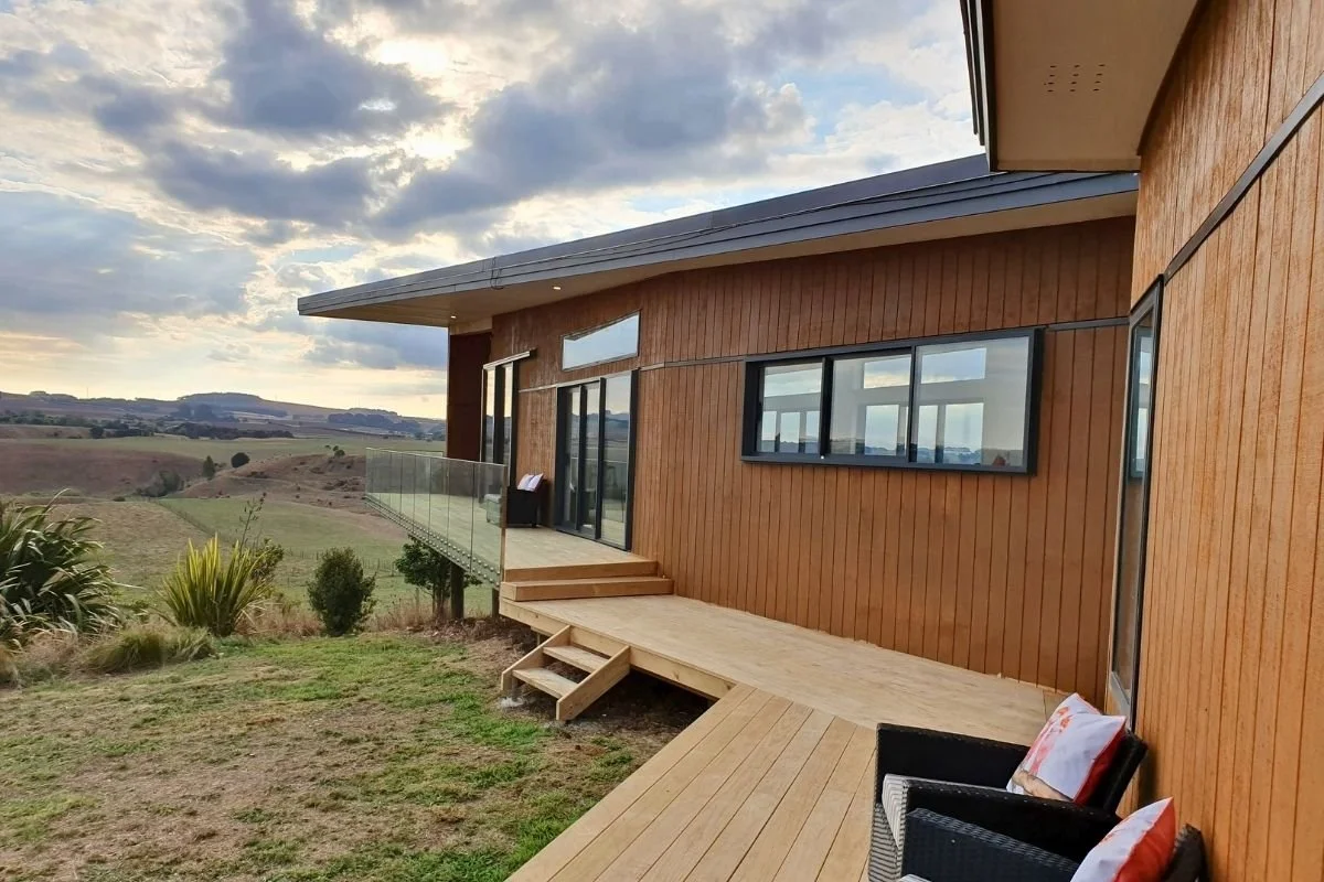 View of a modern house with wooden exterior walls and large windows, situated on a hillside with a deck and outdoor seating area, overlooking rolling countryside and a cloudy sky.