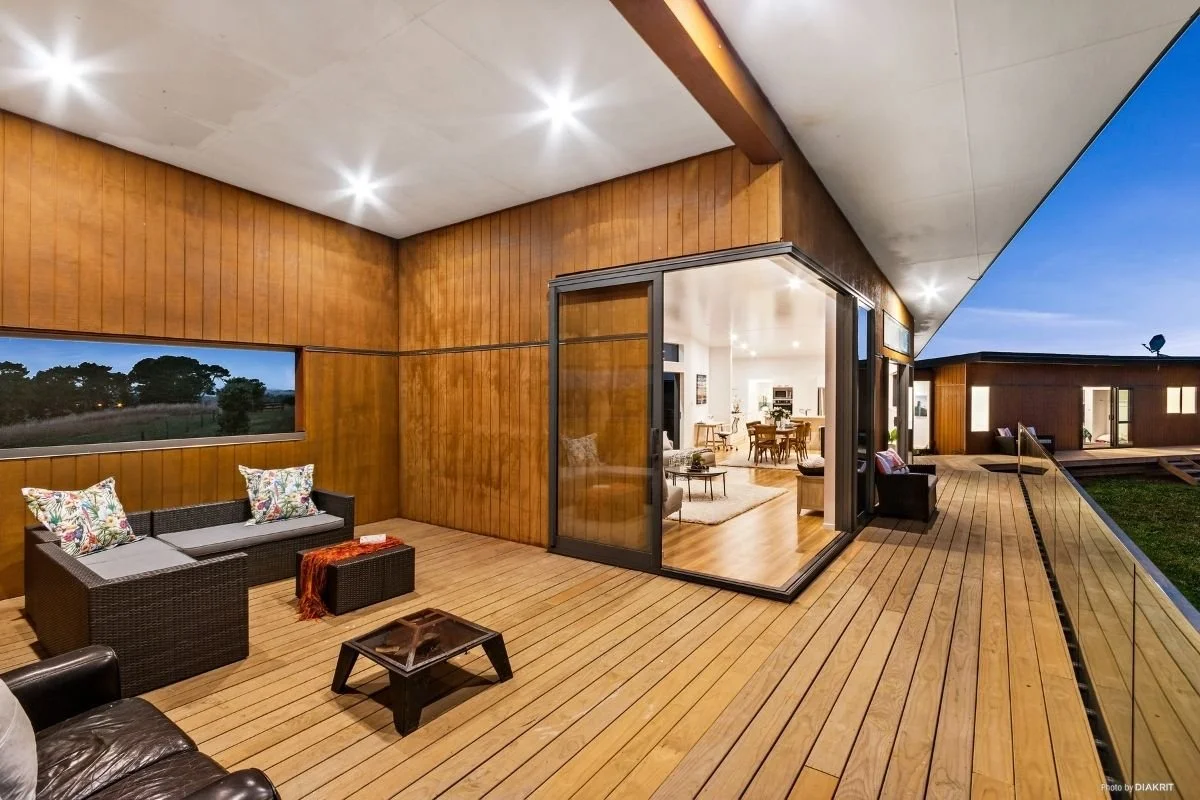 Modern wooden deck with outdoor seating, including a couch with colorful pillows, a small table, and a fire pit, adjoining a house with large glass sliding doors revealing a bright indoor living space at dusk.