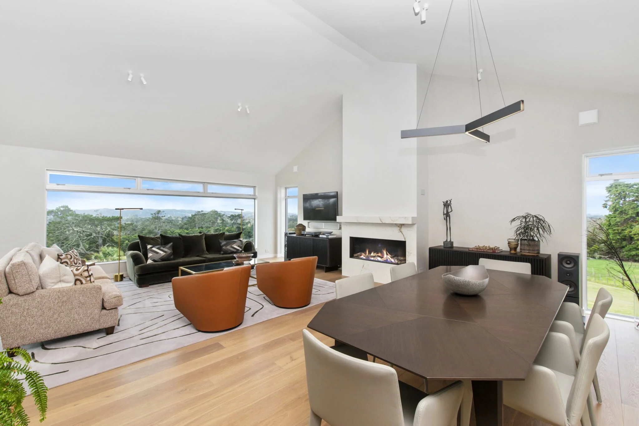 Modern living room with large windows, a gas fireplace, a TV, and contemporary furniture including a beige sofa, black chairs, and a wooden dining table with white chairs.